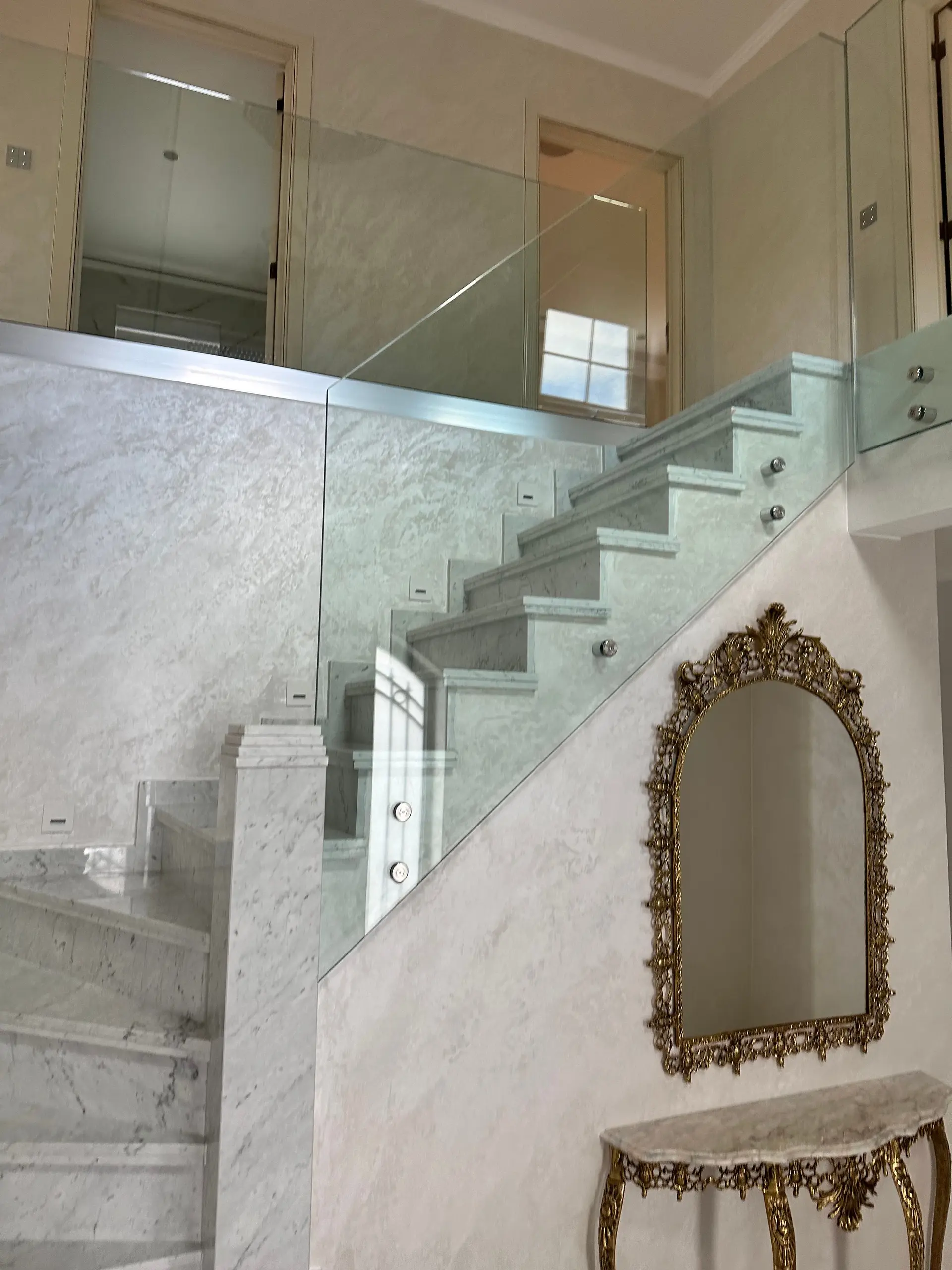 Escalier en marbre terminé avec garde-corps en verre, un miroir doré ouvragé et une console en marbre dans le hall.