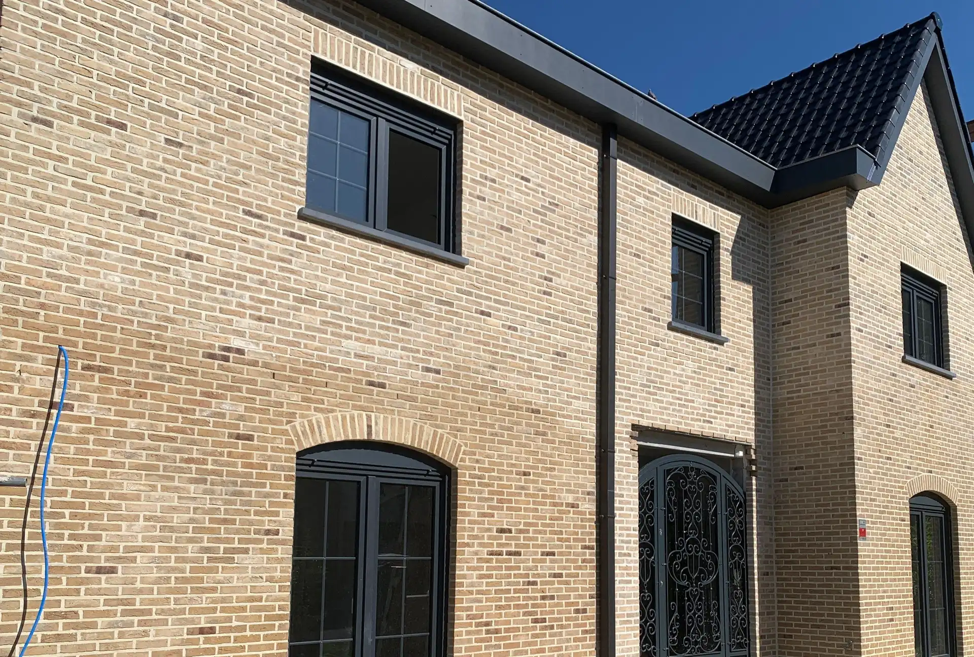 Façade achevée d'une maison de deux niveaux en brique claire, châssis anthracite, porte en fer forgé sous arc de brique et toiture en tuiles noires au soleil