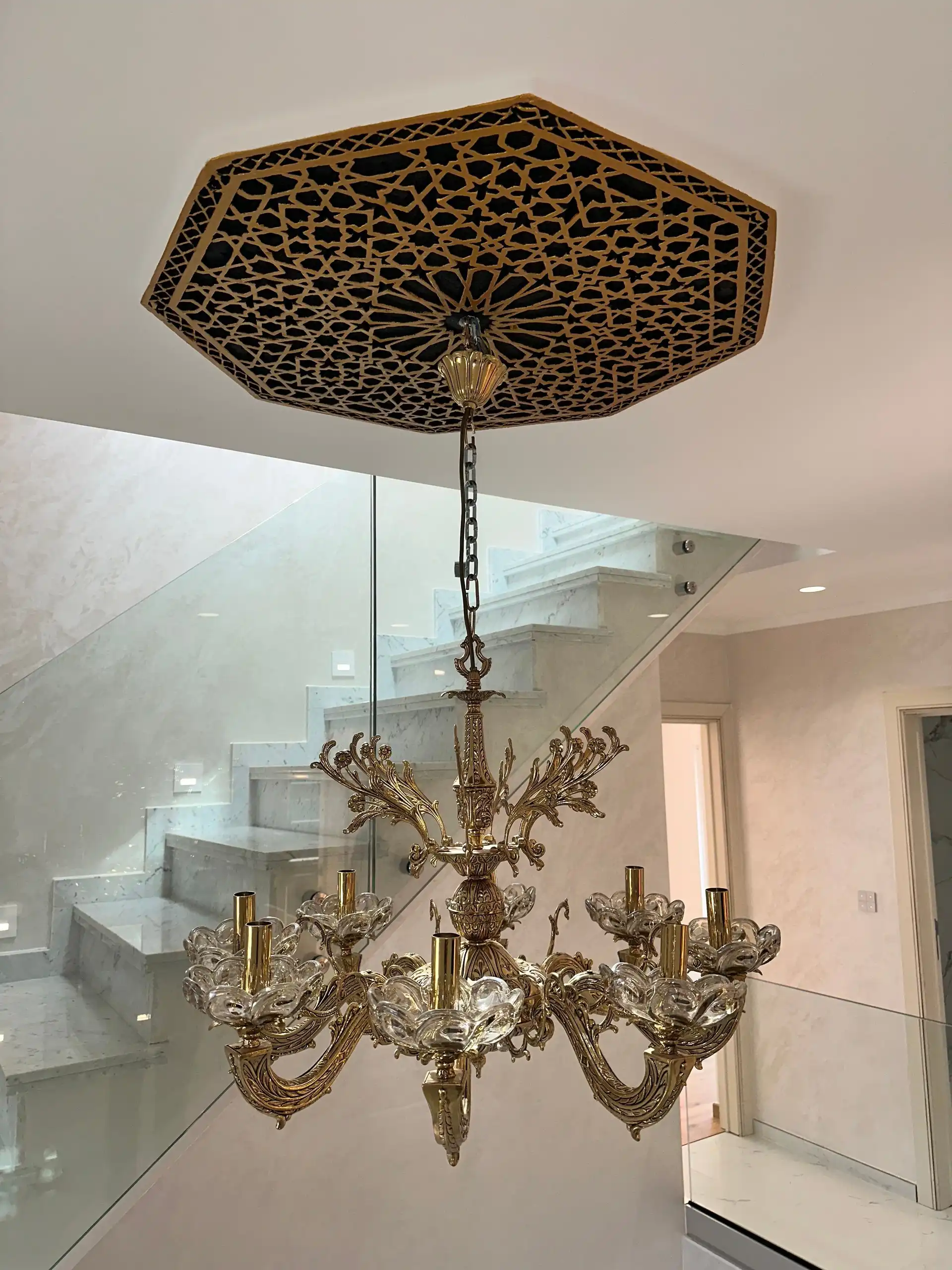 Lustre en cristal suspendu à une rosace de plafond ajourée, avec en arrière-plan l’escalier en marbre et le garde-corps en verre