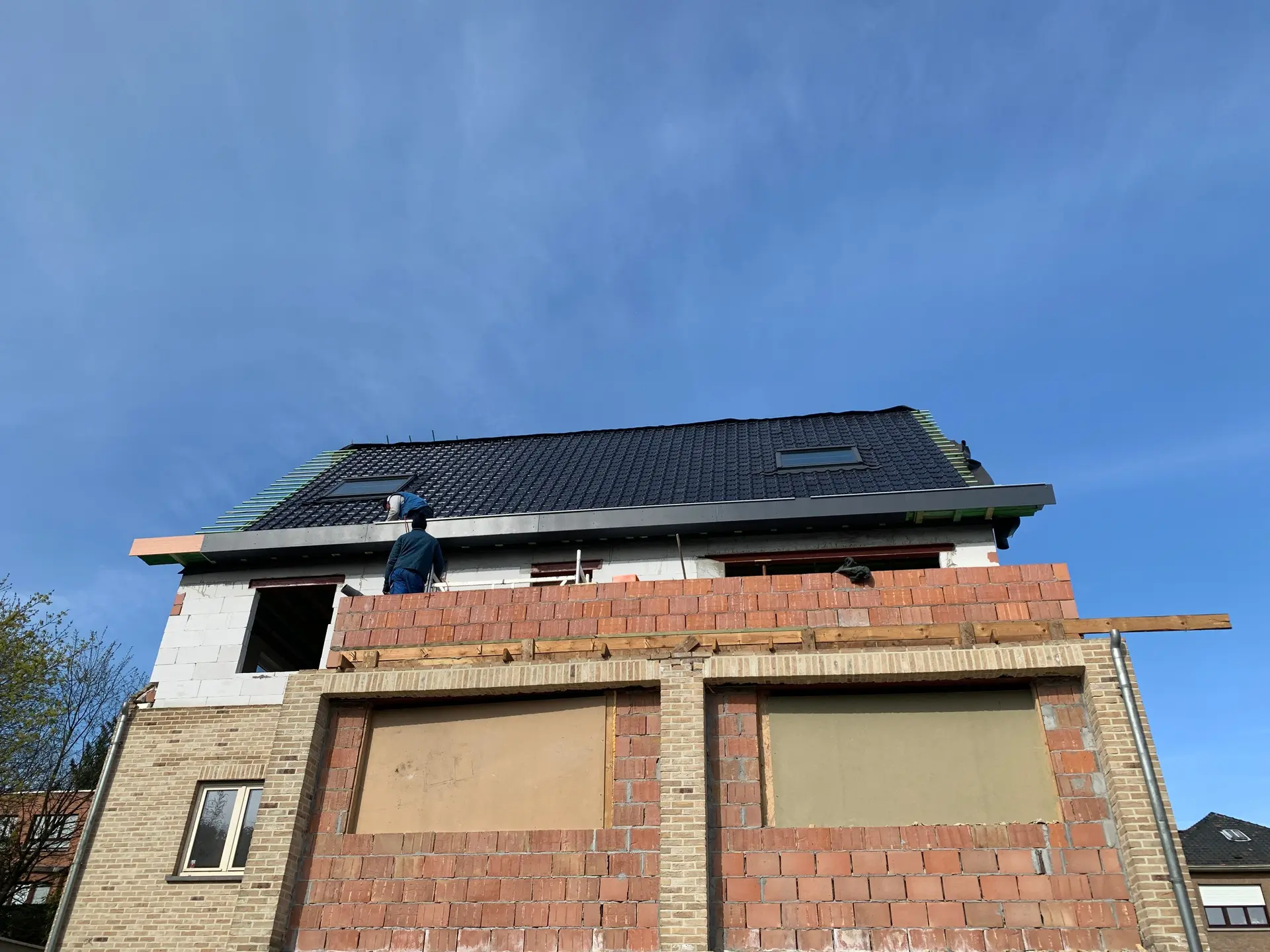 Chantier de la surélévation : le nouvel étage en blocs cellulaires blancs et maçonnerie rouge sous la nouvelle toiture noire, deux couvreurs au travail, grand ciel bleu