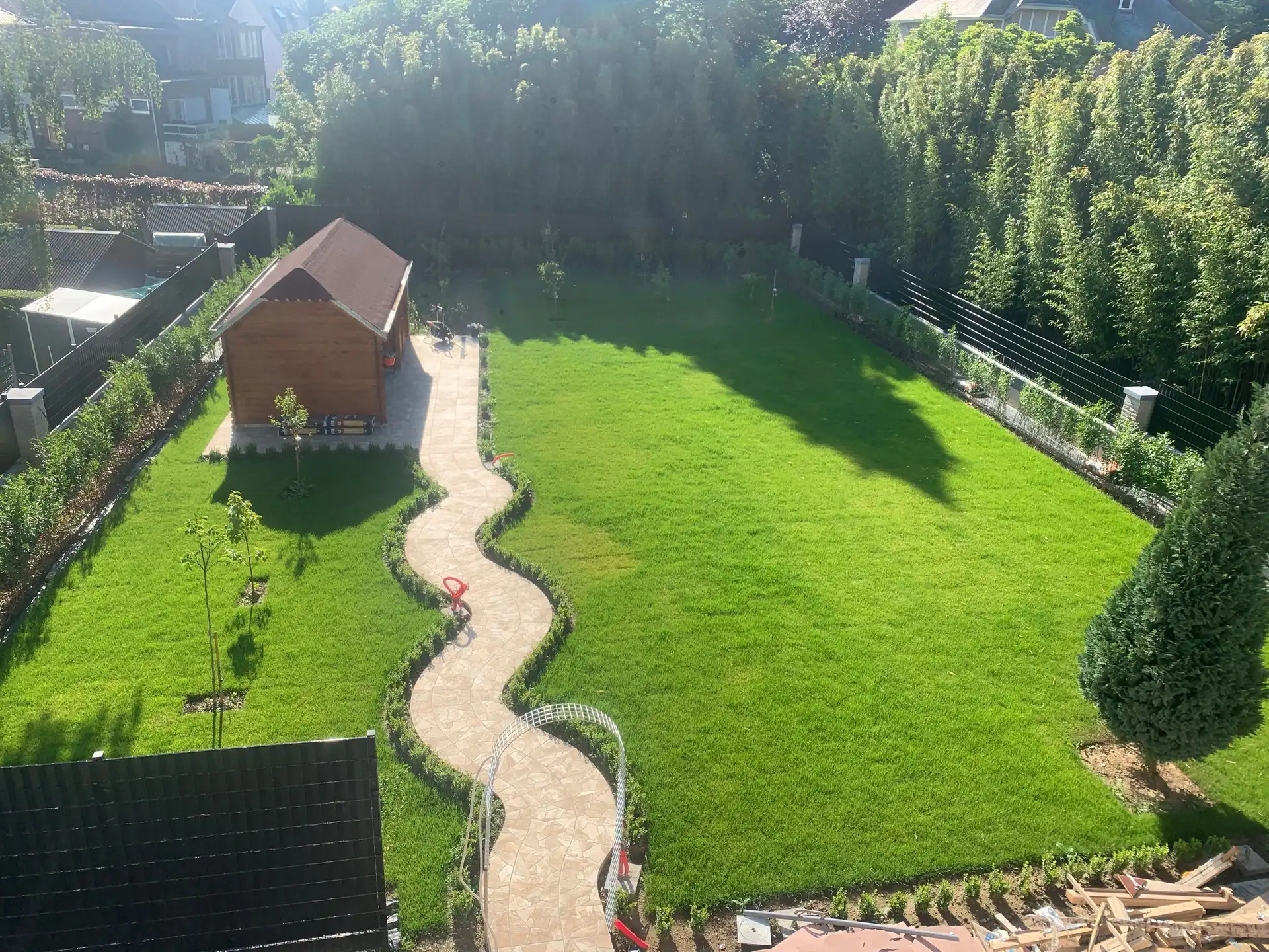Jardin aménagé vu d’en haut : nouvelle pelouse, allée sinueuse en dalles de pierre naturelle, jeunes haies et abri de jardin en bois à toit rouge