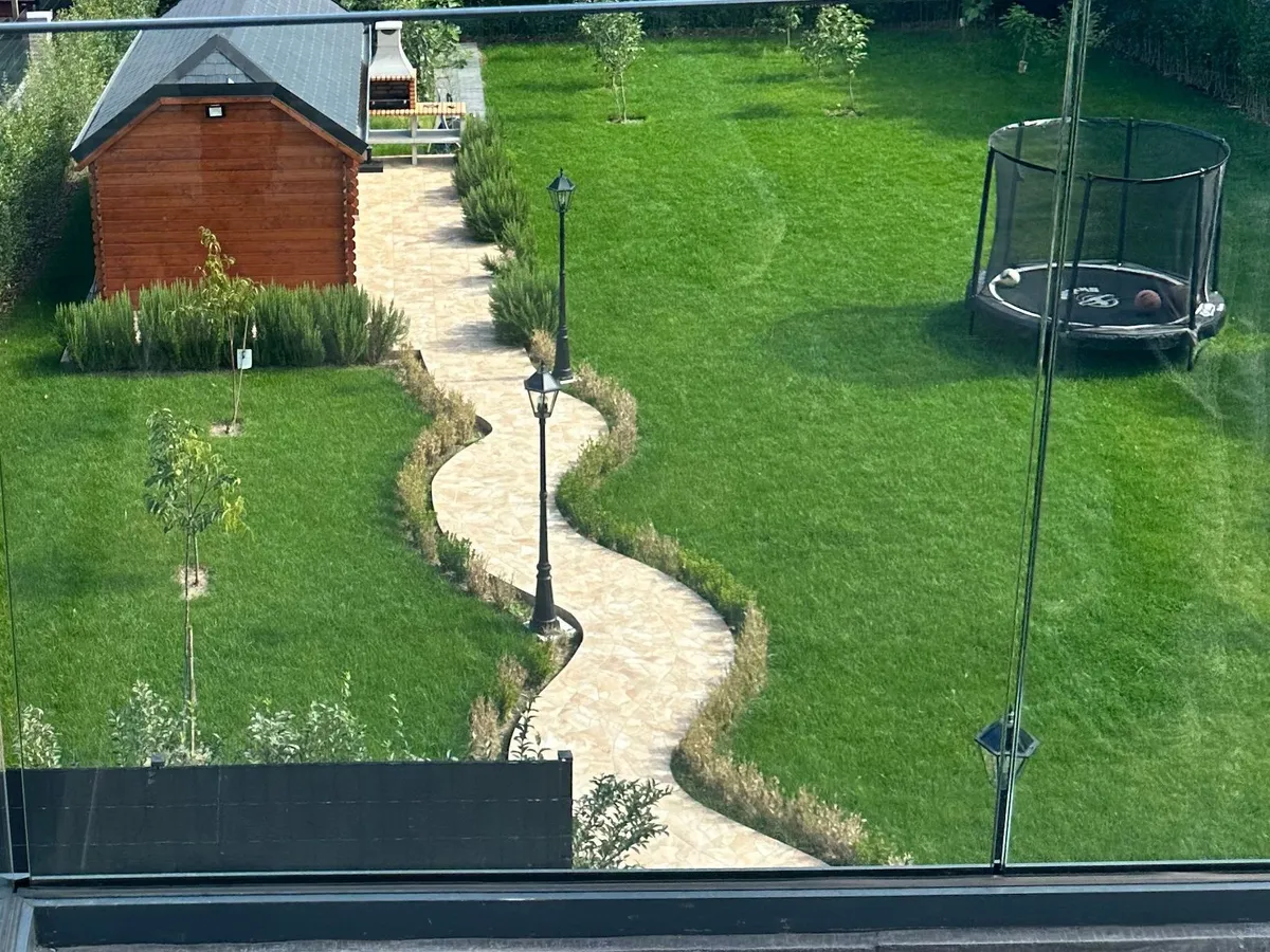 Terrasse achevée avec garde-corps en verre donnant sur le jardin aménagé : pelouse, allée sinueuse en pierre naturelle, haies et abri de jardin en bois
